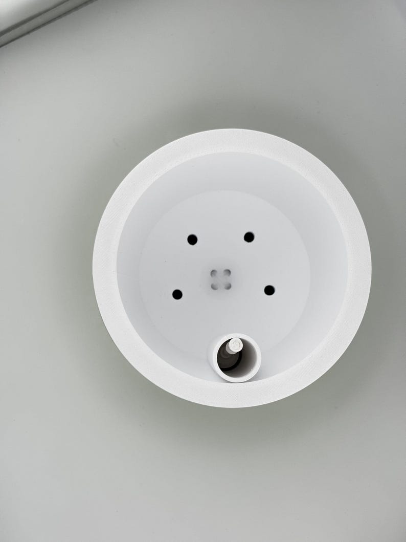 Looking straight down into the white soil insert, showing four small drainage holes, a tiny engraved part code in the centre, and the dark float bead sitting in the water gauge tube at the edge.