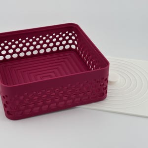 May include: A square magenta storage basket with a perforated lattice pattern on the sides and a stepped square design on the interior base. Beside the basket lies a flat white square lid featuring a series of concentric circular ridges.