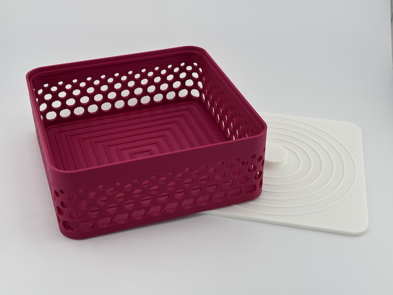 May include: A square magenta storage basket with a perforated lattice pattern on the sides and a stepped square design on the interior base. Beside the basket lies a flat white square lid featuring a series of concentric circular ridges.
