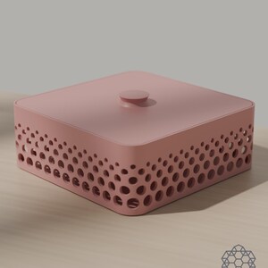 May include: A square, dusty pink storage box with a lid featuring a central circular knob. The sides of the box are decorated with a perforated pattern of varying circular cutouts that create a gradient effect along the perimeter of the container.