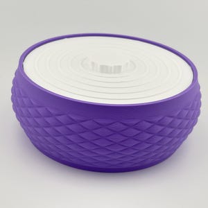 Puede incluir: Un recipiente redondo de color morado con un patr&oacute;n de diamantes texturizado en los lados exteriores. El recipiente cuenta con una tapa circular blanca plana con una serie de anillos conc&eacute;ntricos y un peque&ntilde;o pomo central para levantarla.