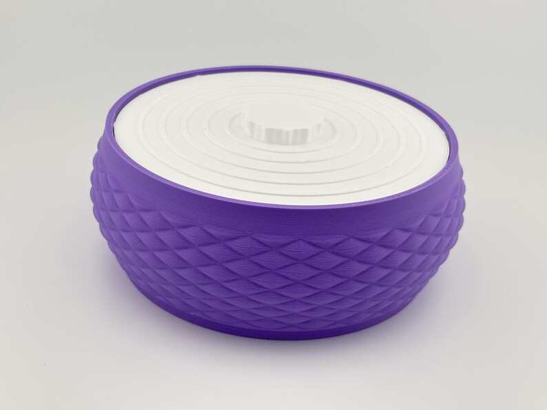 Puede incluir: Un recipiente redondo de color morado con un patr&oacute;n de diamantes texturizado en los lados exteriores. El recipiente cuenta con una tapa circular blanca plana con una serie de anillos conc&eacute;ntricos y un peque&ntilde;o pomo central para levantarla.