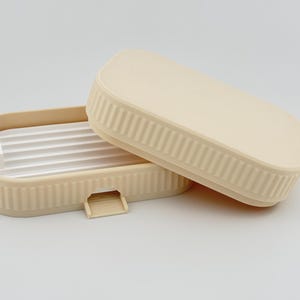 May include: A rectangular beige soap dish with a ribbed exterior and a matching lid. The base features a white, slatted insert for drainage and a small, protruding tab on the front edge. The design is simple, functional, and monochromatic.