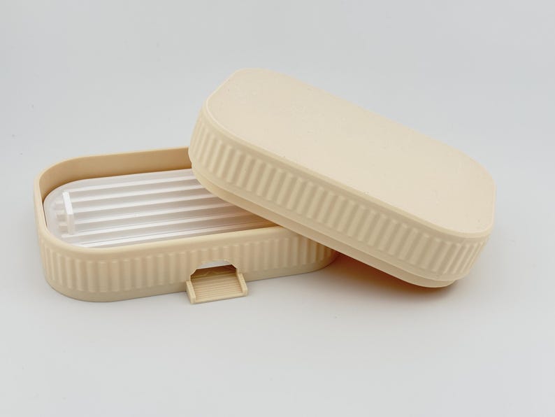 May include: A rectangular beige soap dish with a ribbed exterior and a matching lid. The base features a white, slatted insert for drainage and a small, protruding tab on the front edge. The design is simple, functional, and monochromatic.
