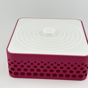 May include: A square storage container with a deep magenta base featuring a perforated pattern of circular cutouts of varying sizes. The container is topped with a flat, white, square lid that has a central circular knob and concentric rings.