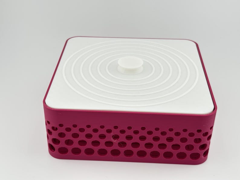 May include: A square storage container with a deep magenta base featuring a perforated pattern of circular cutouts of varying sizes. The container is topped with a flat, white, square lid that has a central circular knob and concentric rings.