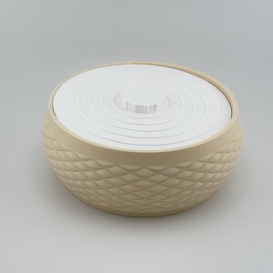 Puede incluir: Un recipiente redondo de color beige con un exterior texturizado de patr&oacute;n de diamantes y una tapa blanca plana con crestas circulares conc&eacute;ntricas. El recipiente tiene una forma ancha y c&oacute;ncava con un pomo central en la tapa.