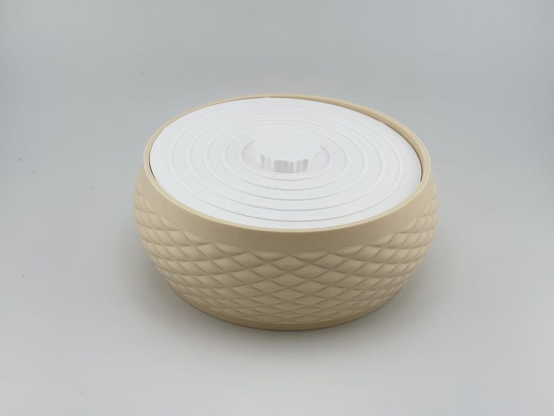 Puede incluir: Un recipiente redondo de color beige con un exterior texturizado de patr&oacute;n de diamantes y una tapa blanca plana con crestas circulares conc&eacute;ntricas. El recipiente tiene una forma ancha y c&oacute;ncava con un pomo central en la tapa.