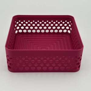 May include: A square magenta storage basket featuring a perforated side wall design with circular cutouts. The interior base of the container displays a series of concentric square indentations, providing a structured and uniform appearance.