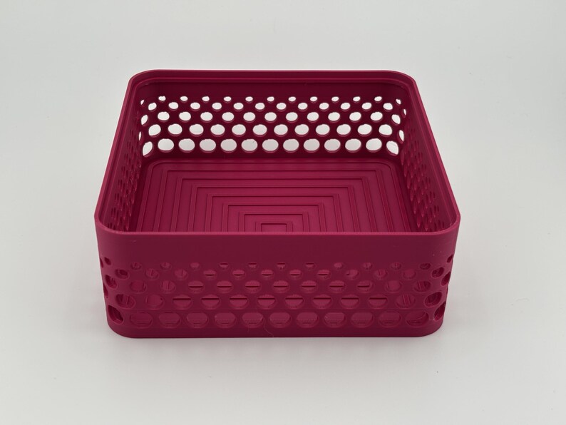 May include: A square magenta storage basket featuring a perforated side wall design with circular cutouts. The interior base of the container displays a series of concentric square indentations, providing a structured and uniform appearance.