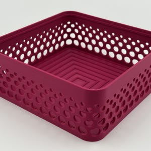 May include: A square magenta storage basket featuring a perforated side pattern with circular cutouts of varying sizes. The interior base of the basket displays a concentric square design, and the overall structure is open-topped and rigid.