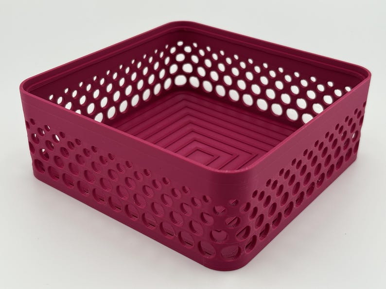 May include: A square magenta storage basket featuring a perforated side pattern with circular cutouts of varying sizes. The interior base of the basket displays a concentric square design, and the overall structure is open-topped and rigid.