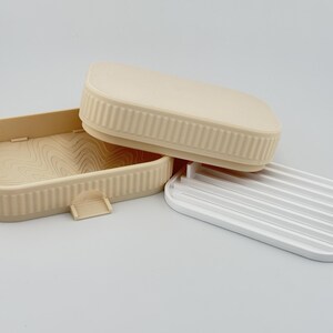 May include: A beige rectangular soap dish with a ribbed exterior and a wavy interior pattern, accompanied by a matching beige lid and a separate white plastic insert featuring parallel drainage slats for holding soap bars securely.