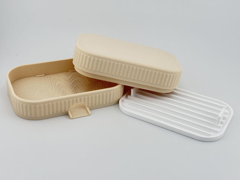 May include: A beige rectangular soap dish with a ribbed exterior and a wavy interior pattern, accompanied by a matching beige lid and a separate white plastic insert featuring parallel drainage slats for holding soap bars securely.