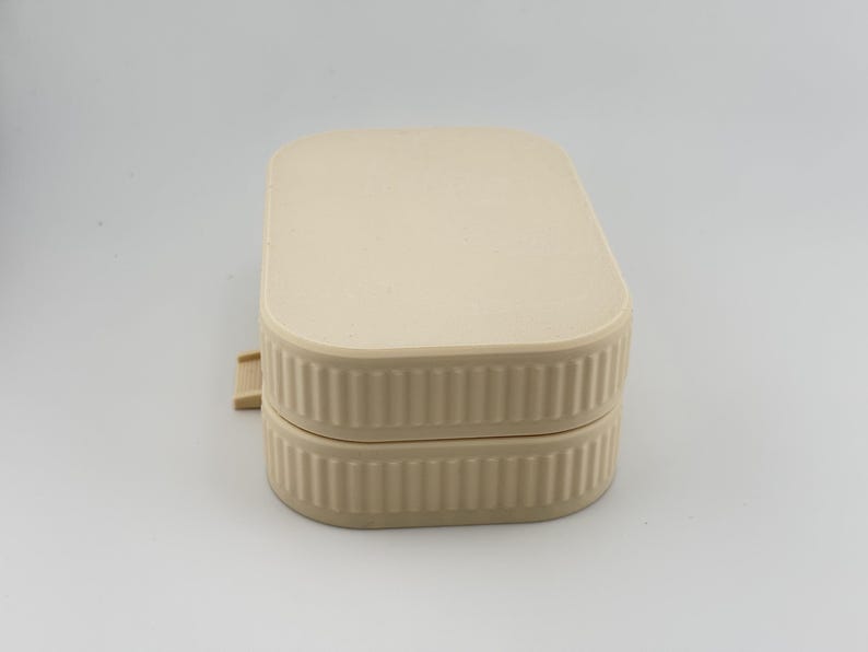 May include: A rectangular, beige-colored storage container with rounded corners and a ribbed, vertical-textured side design. The container features a flat, smooth lid that sits flush with the base, and a small, protruding tab on the left side.