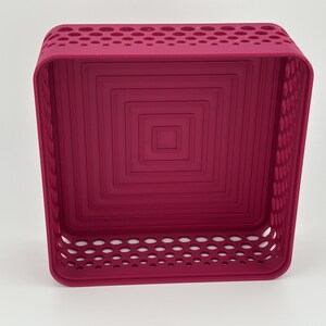May include: A square magenta storage basket featuring a perforated pattern along the upper and lower side walls. The interior base displays a series of concentric square indentations, creating a stepped, layered effect within the container.