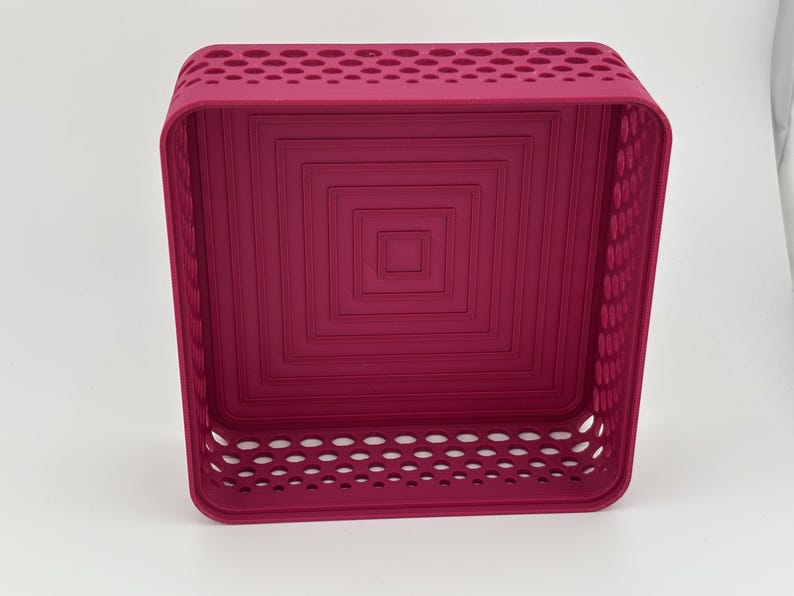 May include: A square magenta storage basket featuring a perforated pattern along the upper and lower side walls. The interior base displays a series of concentric square indentations, creating a stepped, layered effect within the container.