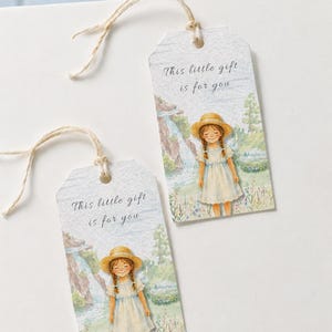 Pode incluir: Duas etiquetas de presente retangulares de papel com cordões claros. Cada etiqueta apresenta uma ilustração em aquarela de uma pessoa com chapéu de palha e vestido claro, em um prado com uma cachoeira. O texto diz, This little gift is for you.
