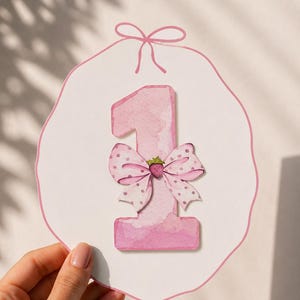 Pode incluir: Um cartão branco recortado com um grande número um em aguarela rosa. O número é decorado com um laço rosa claro às bolinhas e uma pequena ilustração de morango no centro. O cartão tem um contorno rosa e um gráfico de laço no topo.