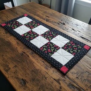 May include: A rectangular quilted table runner featuring a checkerboard pattern of white squares and black squares with a red cherry print. The runner has a black border with a small white cross pattern and four red corner squares with white dots.