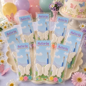 May include: A set of butterfly-shaped plaster figures packaged in clear plastic sleeves with blue sky and cloud backing cards labeled Flutter by Paint your own butterfly. Each kit includes a small paintbrush and three circular paint pots.