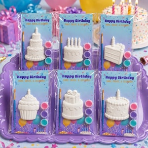 May include: Six individual craft kits in clear plastic packaging, each featuring a white ceramic cake or cupcake shape, a small paintbrush, and three small paint pots. The backing cards display the text Happy Birthday Paint, Create, & Celebrate.