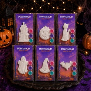 May include: Six individual Halloween-themed paint kits labeled SPOOCTACULAR READY, SET, PAINT. Each kit contains a white ceramic figure—a house, pumpkin, cat, ghost, witch hat, or bat—a small paintbrush, and three circular pots of colored paint.