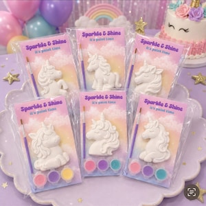 Puede incluir: Seis kits de pintura de unicornio envueltos individualmente en plástico transparente, cada uno con una figura de unicornio de cerámica blanca, un pincel pequeño con mango morado y tres botes de pintura redondos en rosa, morado y turquesa.
