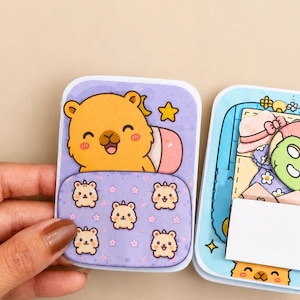 Puede incluir: Un set de manualidades de papel con un personaje de oso de dibujos animados en un saco de dormir morado con un icono de estrella. La sección adyacente muestra varios accesorios de papel en miniatura, incluyendo un sombrero, un vestido floral y una máscara facial verde.