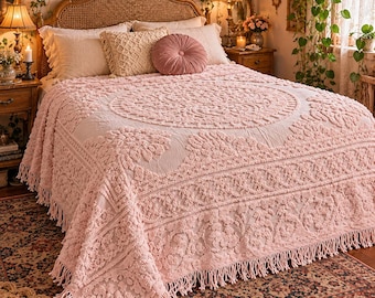 Puede incluir: Una colcha de color rosa claro con un patrón floral en relieve y un borde con flecos. El tejido rectangular cubre una cama y muestra un motivo circular central rodeado de texturas botánicas intrincadas y borlas largas.