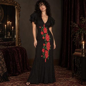 May include: A floor-length black dress featuring short ruffled sleeves, a V-neckline, and a vertical embroidered pattern of vibrant red roses with green leaves cascading down the center of the bodice and skirt, creating a fitted silhouette.