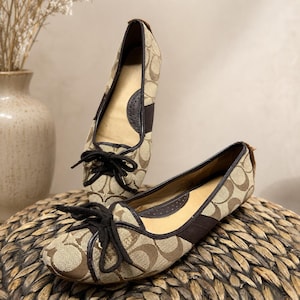 Puede incluir: Un par de bailarinas de lona en color beige y marrón con un patrón de logotipo repetitivo, ribete de cuero marrón oscuro y un lazo negro decorativo en la punta redondeada. El interior está forrado con material beige claro y detalles oscuros.