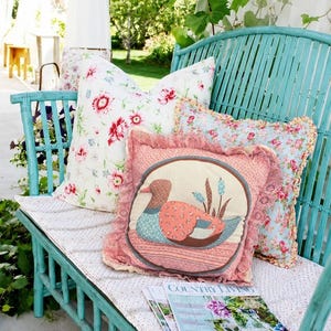 May include: A turquoise slatted bench featuring three decorative pillows. One pillow has a white background with red floral patterns, another has a blue background with floral prints, and the third is a pink square pillow with a duck applique.