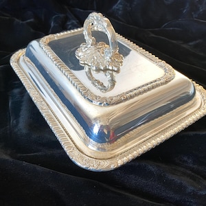 May include: A rectangular silver-plated serving dish with a removable lid. The lid features a decorative embossed border and a central ornate handle with intricate floral detailing. The polished metal surface reflects light across its smooth top.