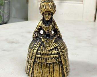 May include: A vintage brass bell shaped like a woman in a long dress and hat. The bell has intricate details and a golden hue. The bell is likely a decorative item or collectible.