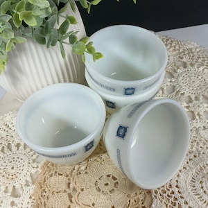 May include: A set of four white, round bowls with a blue geometric design around the rim. The bowls are stacked and arranged on a lace doily. The bowls are made of a thick, opaque material.