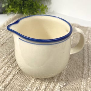 May include: Cream-colored ceramic pitcher with a blue rim and handle. The pitcher has a spout for easy pouring and a simple, elegant design. A thin blue line accents the body of the pitcher. The pitcher is likely used for serving liquids.