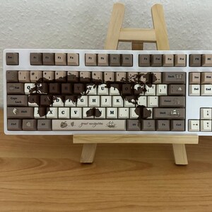 Könnte beinhalten: Eine mechanische Tastatur in voller Größe mit weißem Rahmen und einer Mischung aus hell- und dunkelbraunen Tastenkappen. Die mittleren Tasten zeigen eine dunkelbraune Weltkarte, den Schriftzug "great navigation" und ein kleines Schiff.