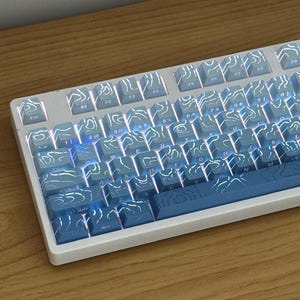 May include: A mechanical keyboard with a white frame and blue keycaps featuring a white topographic map pattern. The keys are backlit with a soft white light, and the layout includes standard alphanumeric characters, function keys, and a spacebar.
