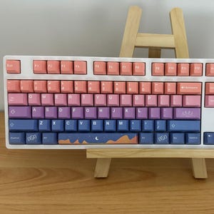 Könnte beinhalten: Eine mechanische Tastatur in voller Größe mit weißem Rahmen und einem Tastenkappen-Set im Farbverlauf, das von Pfirsich oben zu Rosa, Lila und Dunkelblau unten übergeht. Die Leertaste zeigt eine Grafik mit Mondsichel und Sternen.