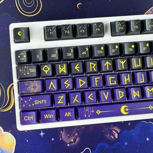 Könnte beinhalten: Eine weiße mechanische Tastatur mit dunkelvioletten Tastenkappen, die mit gelben Himmels- und Runensymbolen verziert sind. Die Tasten zeigen Mondphasen, Sterne und geometrische Muster neben den üblichen alphanumerischen Zeichen.