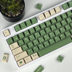 Op de afbeelding: Een mechanisch toetsenbord met een wit frame en een mix van saliegroene en crèmekleurige toetsen. De toetsen bevatten Engelse letters en Japanse hiragana-tekens. Sommige toetsen tonen iconen van bladeren en een theepot, met een losse toets in het Japans.