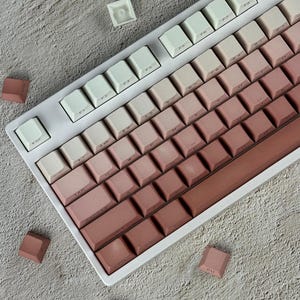 Könnte beinhalten: Eine mechanische Tastatur mit einem Farbverlauf-Tastenkappenset, das von hellem Creme oben links zu einem tiefen Altrosa unten rechts übergeht. Die Tastatur hat einen weißen Rahmen und mehrere lose, nicht angebrachte Tastenkappen liegen verstreut daneben.