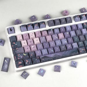 May include: A mechanical keyboard with a white frame and custom keycaps in shades of purple and dark blue. The keys feature floral patterns, butterflies, stars, a crescent moon, and a black cat icon. Several loose keycaps are scattered nearby.
