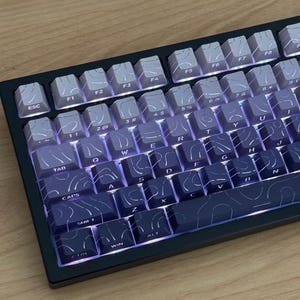 Könnte beinhalten: Eine mechanische Tastatur mit einem Farbverlauf von Hellgrau oben bis Dunkelmarineblau unten. Jede Tastenkappe zeigt ein weißes topografisches Linienmuster und eine hintergrundbeleuchtete Beschriftung, eingefasst in einen schwarzen Rahmen.