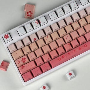 Könnte beinhalten: Eine mechanische Tastatur mit weißem Rahmen und einem Farbverlauf aus rosa Tastenkappen. Die Tasten zeigen Kirschblütenmotive und Standardzeichen. Mehrere lose Tastenkappen mit Blumenmustern sind um die Tastatur herum verstreut.