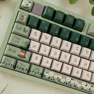 Può includere: Una tastiera meccanica con telaio verde salvia e un set di tasti personalizzato. I tasti presentano una combinazione di rosa chiaro, verde scuro e verde salvia, decorati con illustrazioni botaniche di piante in vaso, viti e fiori bianchi.