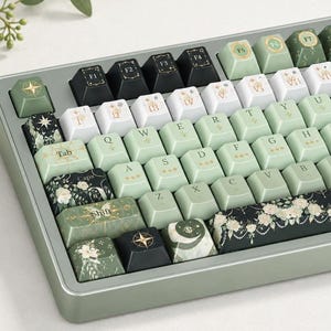 May include: A mechanical keyboard featuring a sage green frame and a custom keycap set. The keys display a mix of light green, white, and black colors, decorated with floral patterns, celestial motifs, and Roman numerals on the function row.