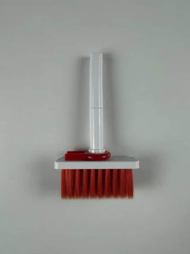 May include: A cleaning brush featuring a white plastic handle and a rectangular white base. The brush head is equipped with dense, straight red bristles. A red plastic connector piece attaches the handle to the base of the brush head.