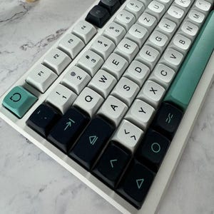 Può includere: Una tastiera meccanica con telaio bianco e un mix di tasti bianchi, neri e verde acqua. I tasti bianchi mostrano caratteri alfanumerici e simboli neri. I tasti neri presentano simboli verde acqua, mentre i tasti verde acqua includono un'icona a freccia circolare e una lunga barra spaziatrice. Il layout segue una configurazione QWERTY con tasti funzione lungo la riga superiore.
